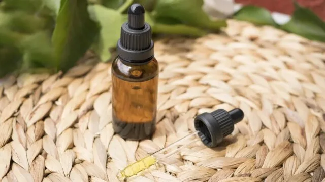 An amber glass dropper bottle with a pipette containing herbal fluid extract, placed on a woven mat with green leaves in the background — representing a homemade herbal tincture.