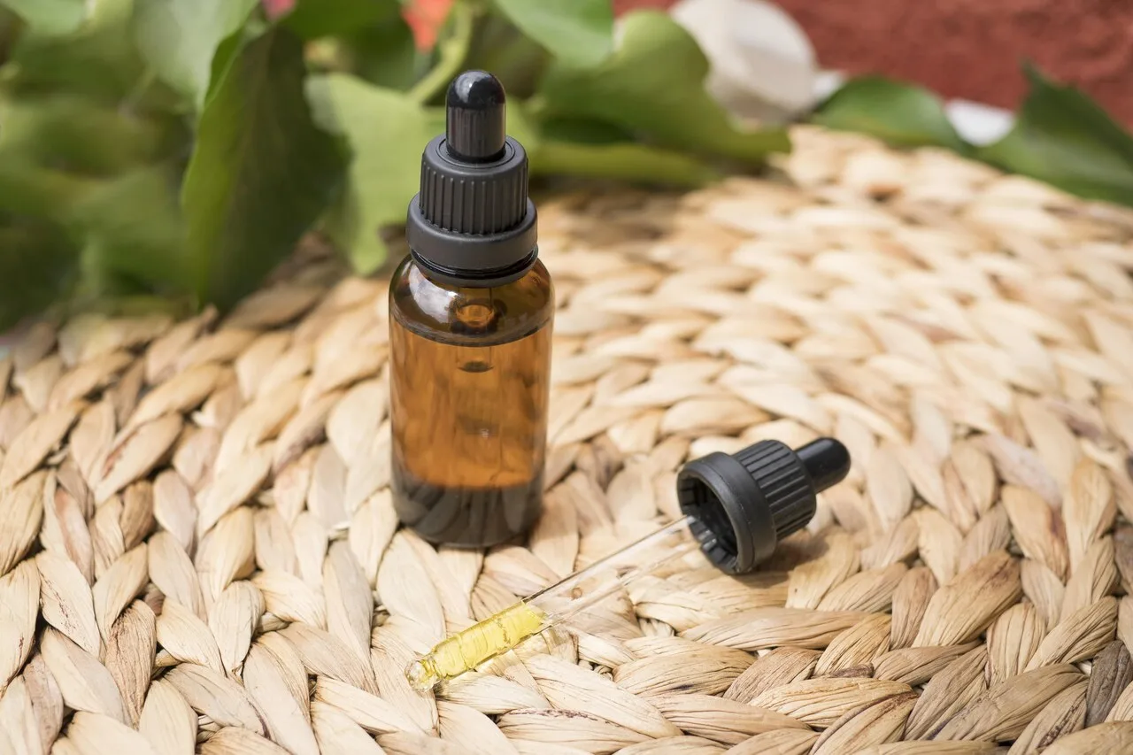 An amber glass dropper bottle with a pipette containing herbal fluid extract, placed on a woven mat with green leaves in the background — representing a homemade herbal tincture.