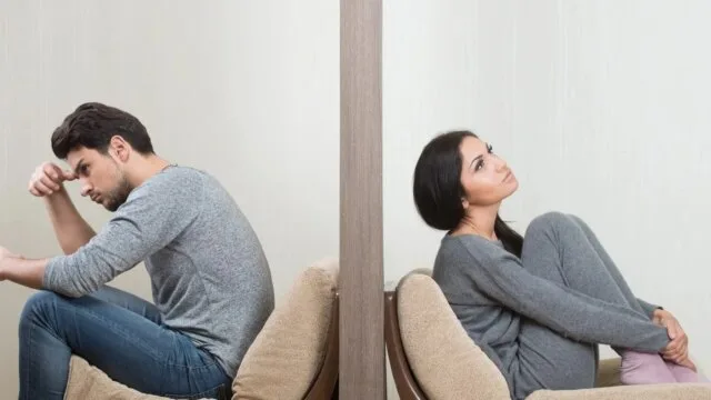 A man and a woman sitting back-to-back on separate armchairs, divided by a wall, both looking away — illustrating emotional distance and avoidant attachment in adult relationships.