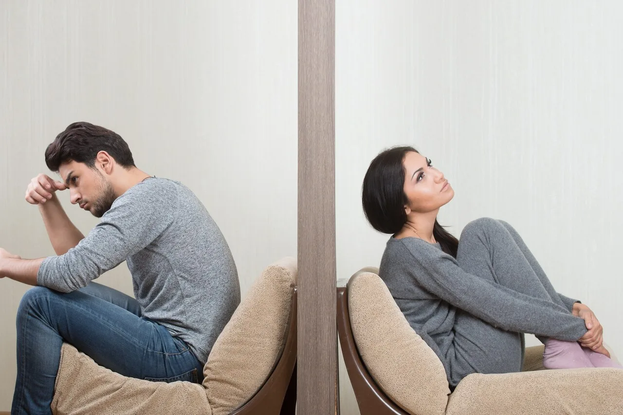 A man and a woman sitting back-to-back on separate armchairs, divided by a wall, both looking away — illustrating emotional distance and avoidant attachment in adult relationships.