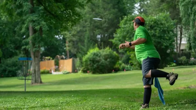 A disc golf player releasing a disc mid-throw toward a basket target on a green outdoor course — illustrating the rotational mechanics and technique essential for improving disc golf performance.