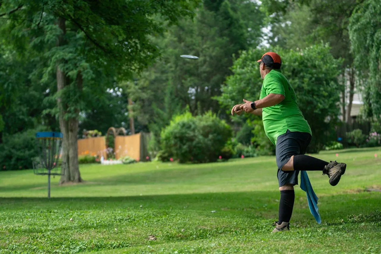 A disc golf player releasing a disc mid-throw toward a basket target on a green outdoor course — illustrating the rotational mechanics and technique essential for improving disc golf performance.