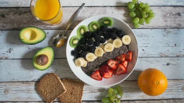 A bowl of high-fiber foods including berries, banana, chia seeds and kiwi surrounded by avocado, whole grain bread and fresh fruit