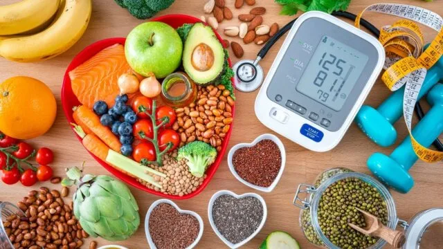 Healthy foods including fruits, vegetables, nuts and salmon arranged on a heart-shaped plate alongside a blood pressure monitor and dumbbells