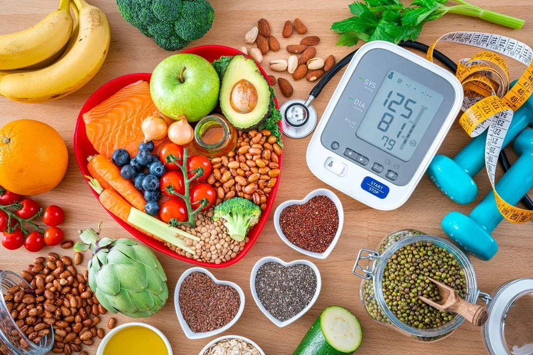 Healthy foods including fruits, vegetables, nuts and salmon arranged on a heart-shaped plate alongside a blood pressure monitor and dumbbells