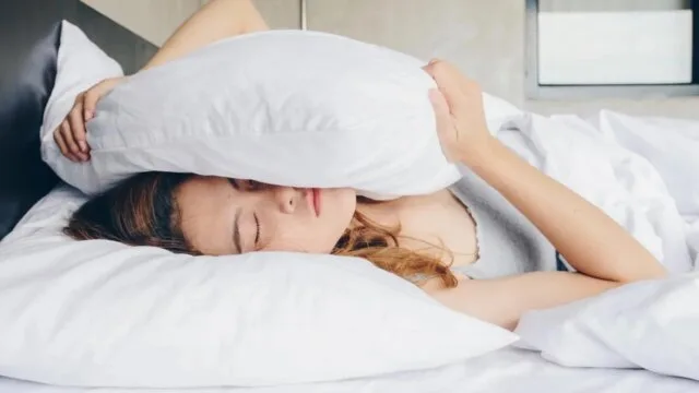 A woman lying in bed covering her head with a pillow, struggling with sleeplessness and anxiety — illustrating the cycle of worry and restless nights.