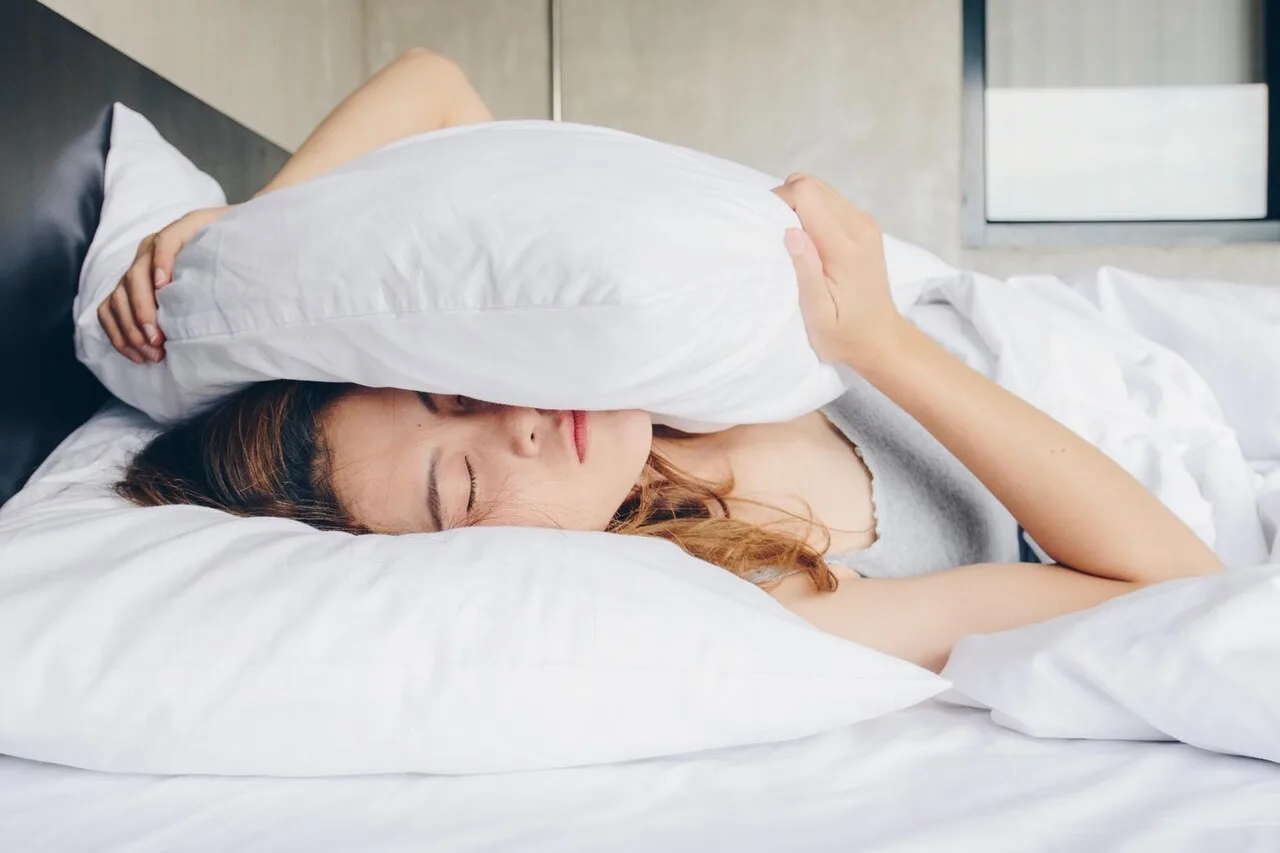 A woman lying in bed covering her head with a pillow, struggling with sleeplessness and anxiety — illustrating the cycle of worry and restless nights.