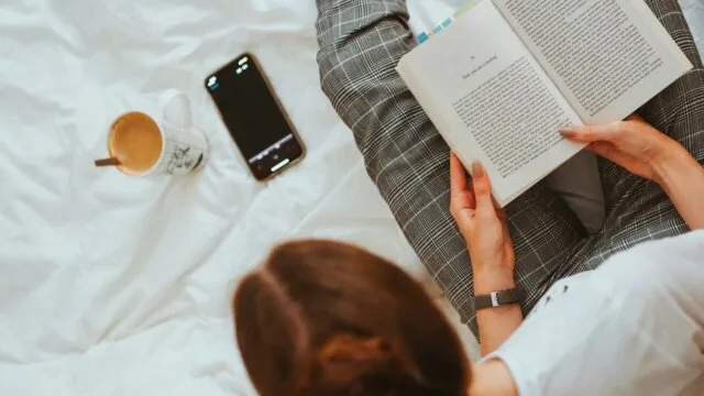 A woman reading a physical book in bed with her phone set aside and a cup of coffee nearby — representing a mindful break from screens to improve digital wellbeing.