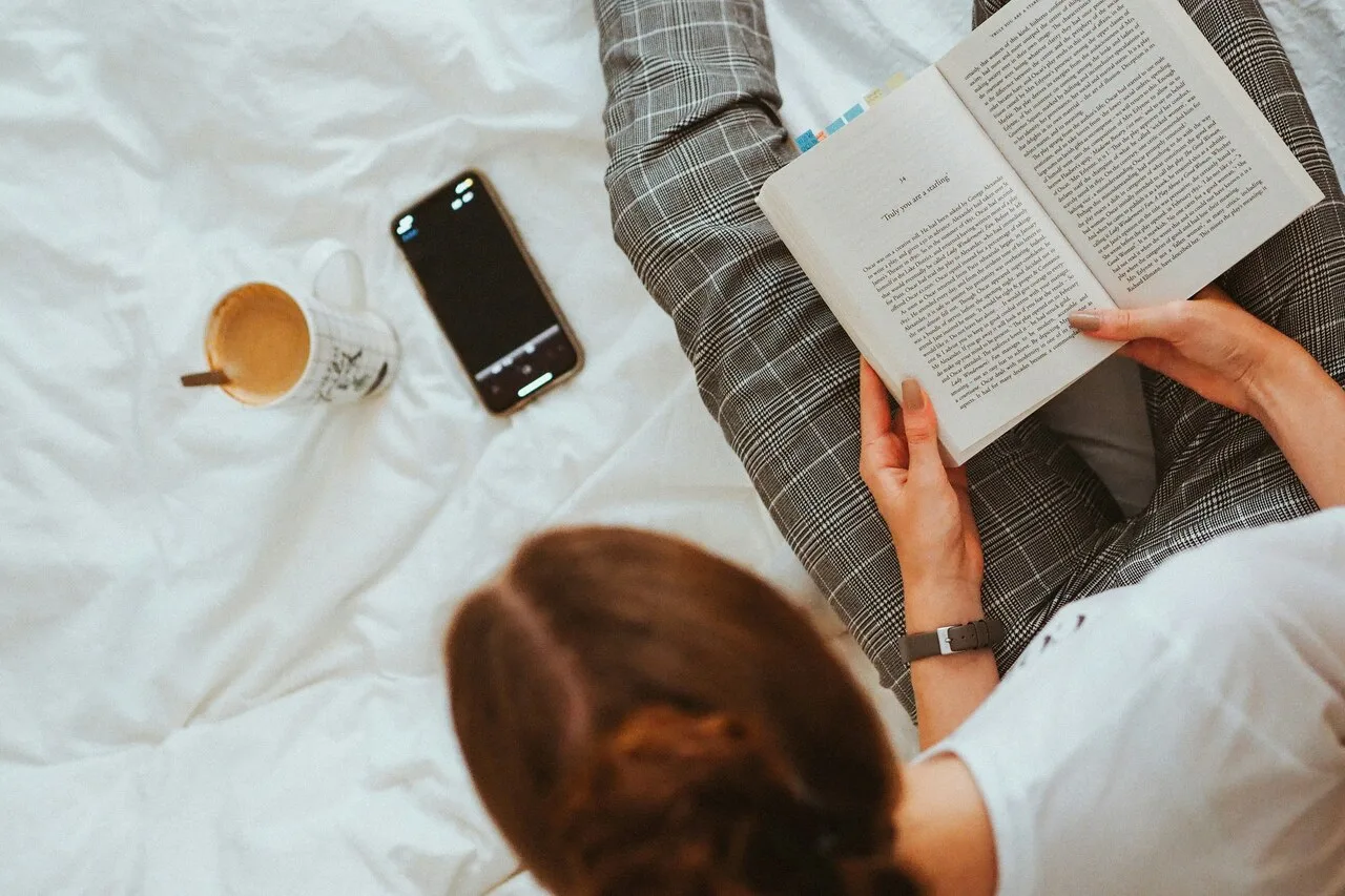 A woman reading a physical book in bed with her phone set aside and a cup of coffee nearby — representing a mindful break from screens to improve digital wellbeing.