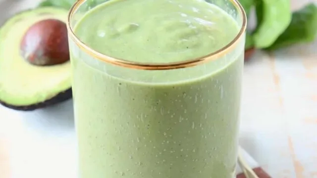 Green superfood smoothie in a glass with a spoon of greens powder, fresh spinach and avocado in the background representing the best greens powder supplement