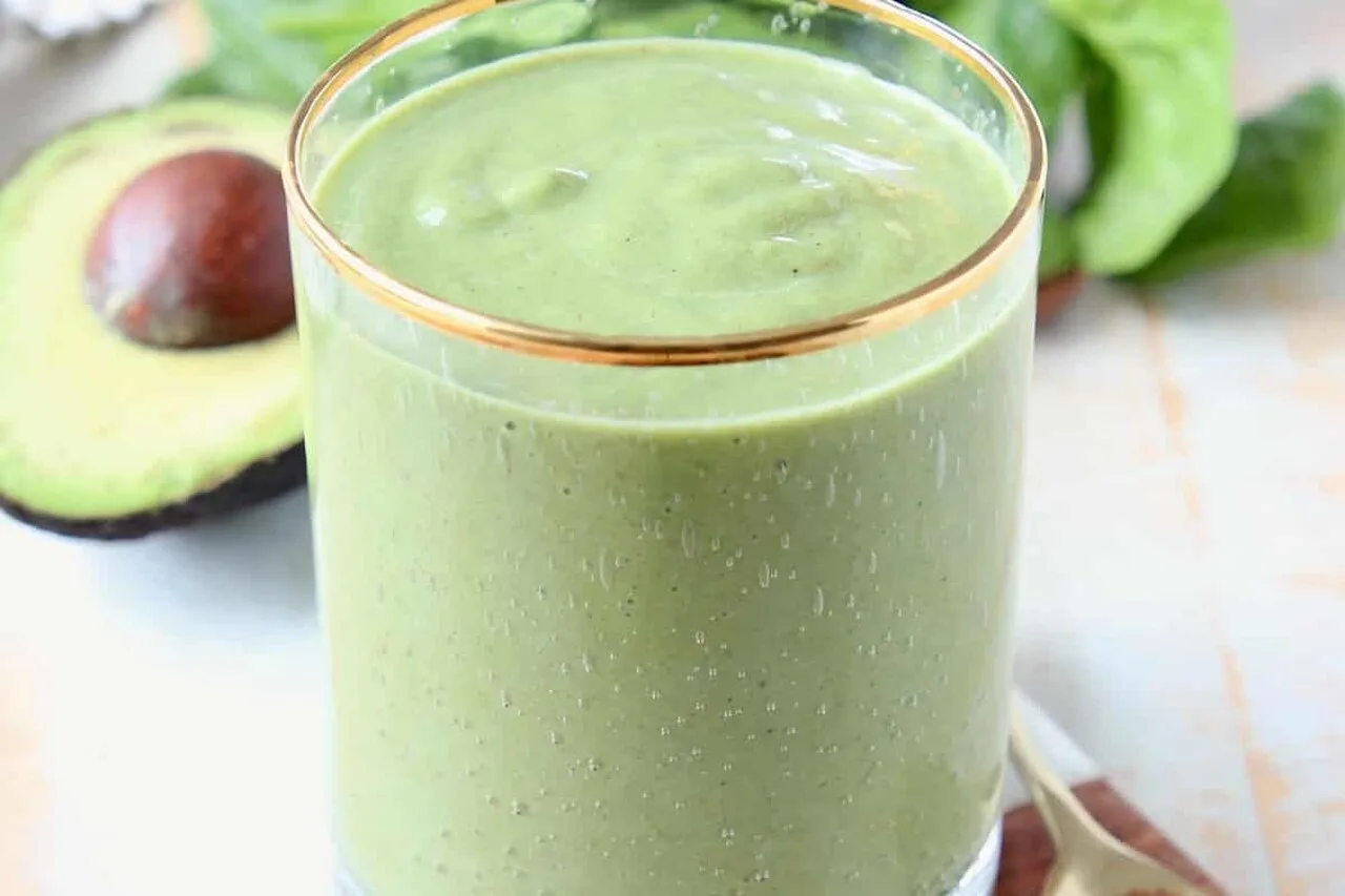 Green superfood smoothie in a glass with a spoon of greens powder, fresh spinach and avocado in the background representing the best greens powder supplement