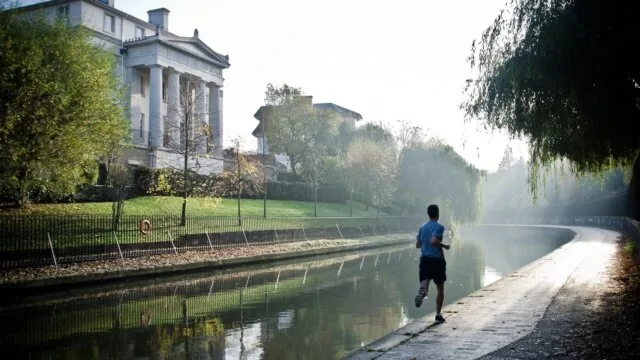 A man jogging along a misty canal path in the early morning surrounded by trees and historic buildings β representing the consistency and dedication essential for long-term fitness progress.