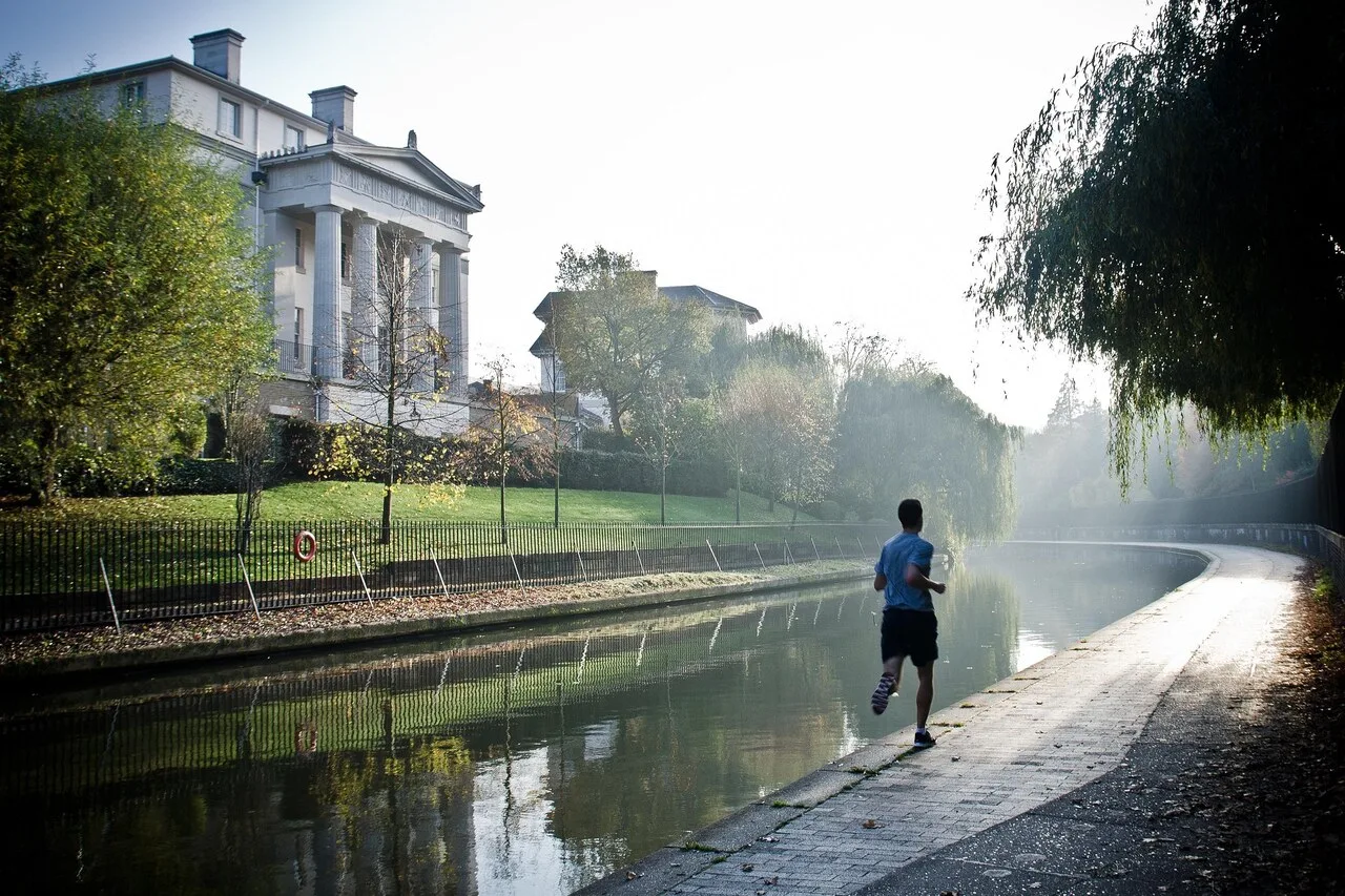 A man jogging along a misty canal path in the early morning surrounded by trees and historic buildings β representing the consistency and dedication essential for long-term fitness progress.