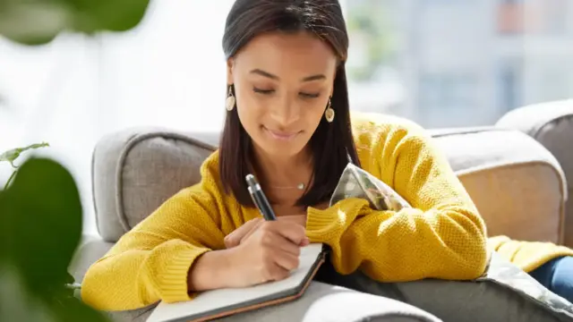 Young woman smiling while writing in a self-compassion journal on a sofa, practicing mindfulness and emotional resilience at home
