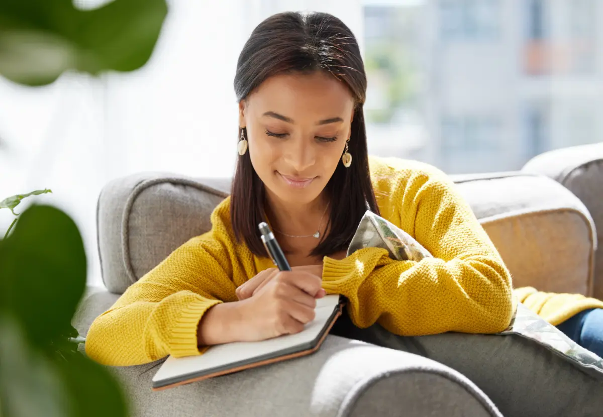Young woman smiling while writing in a self-compassion journal on a sofa, practicing mindfulness and emotional resilience at home