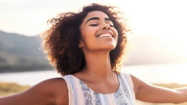 Young woman with arms open and eyes closed smiling peacefully outdoors, practicing mindfulness and body gratitude in nature