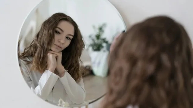 Young woman looking at herself in a round mirror practicing self-awareness to overcome people-pleasing