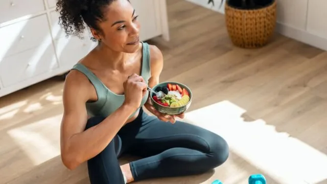 Fit woman eating a healthy fruit bowl after exercise with dumbbells nearby representing balanced nutrition and fitness as sustainable health optimization strategies for longevity