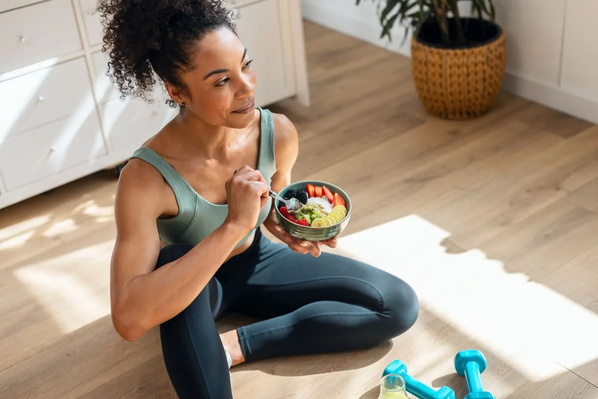 Fit woman eating a healthy fruit bowl after exercise with dumbbells nearby representing balanced nutrition and fitness as sustainable health optimization strategies for longevity