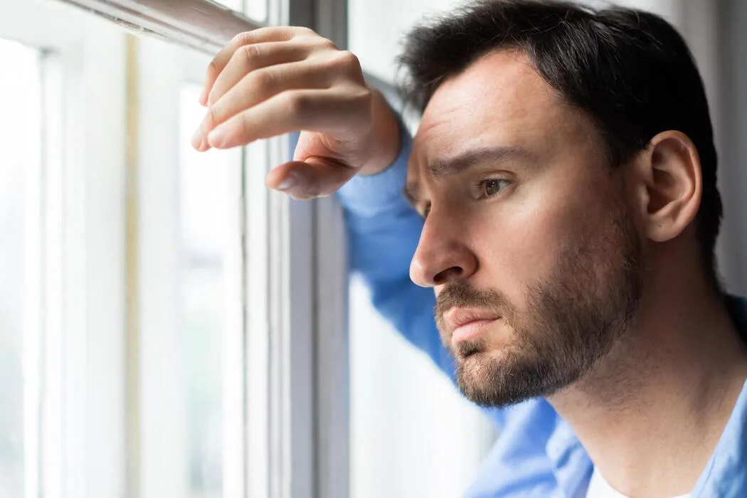 Man gazing thoughtfully out a window representing emotional suppression and the hidden cost of saying I'm fine